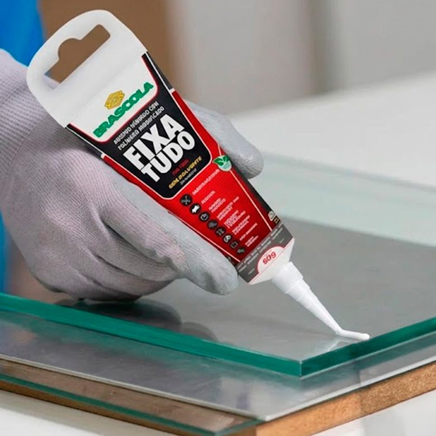 Mão com luva aplicando filete de adesivo Fixa Tudo Brascola branco sobre placa de vidro transparente