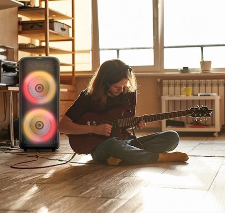 Homem tocando guitarra conectado à Caixa de Som Philips Party Speaker em ambiente iluminado