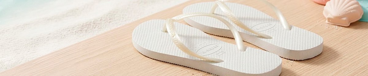 Havaianas Kids Slim brancas sobre madeira com fundo de areia e conchas, cenário de praia.