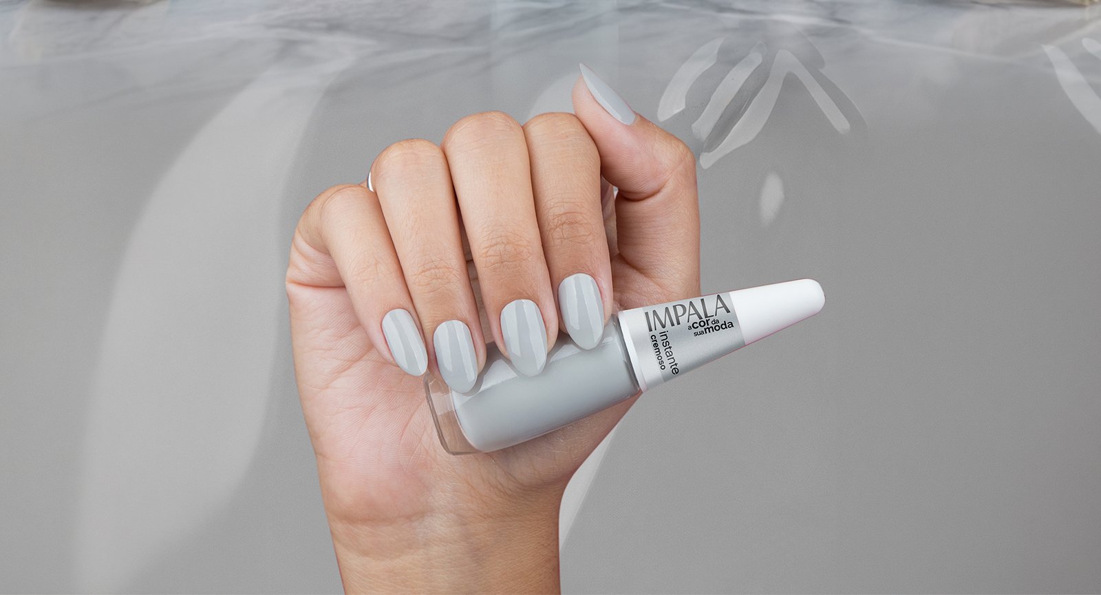 Mão segurando Esmalte Impala Instante cinza claro cremoso aplicado em unhas com fundo cinza